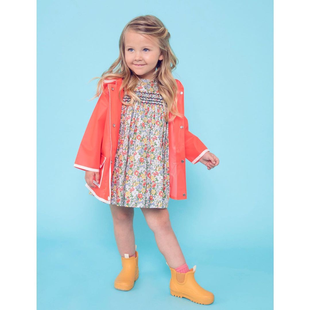 A young girl poses in a floral dress, orange raincoat with white trim, and matching yellow rainboots against a light blue backdrop.