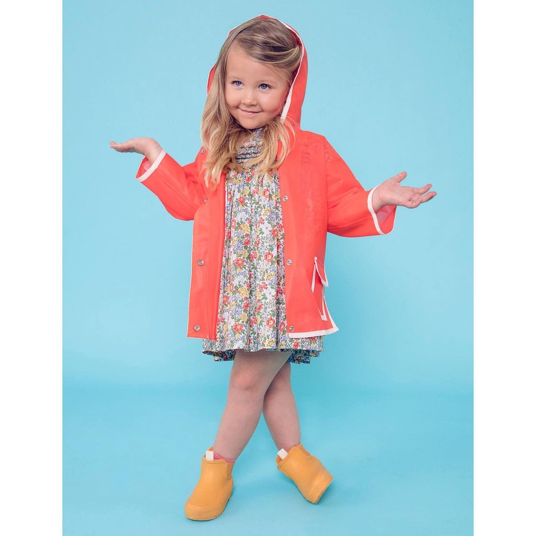 Young girl in a coral raincoat and yellow boots smiles with arms outstretched against a light blue backdrop in a full-body shot.