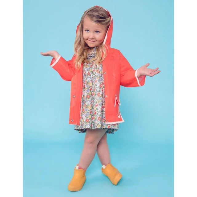 Young girl in a coral raincoat and yellow boots smiles with arms outstretched against a light blue backdrop in a full-body shot.