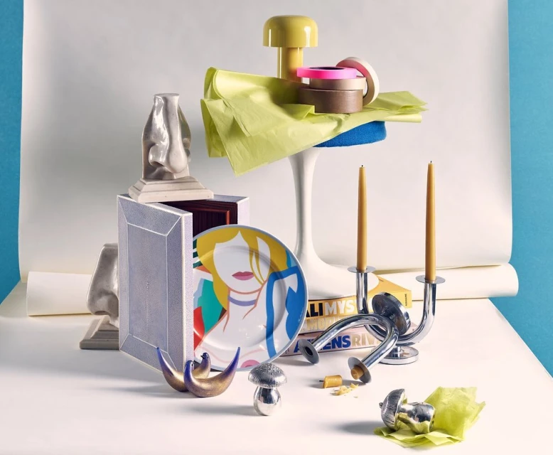 Still life of decorative objects in bright lighting features candlesticks, vases, book, mushroom sculpture, candle, and geometric shapes.