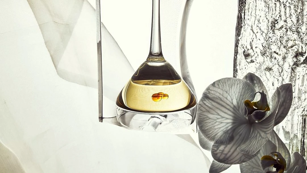 Still life shot of an elegant perfume bottle with golden liquid set against a bright backdrop with a white orchid in the foreground.