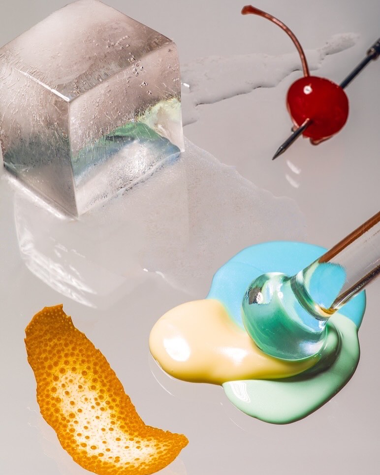 Studio shot of a melting ice cube, cherry, citrus peel, and colorful liquid for a food and beverage concept with bright lighting.