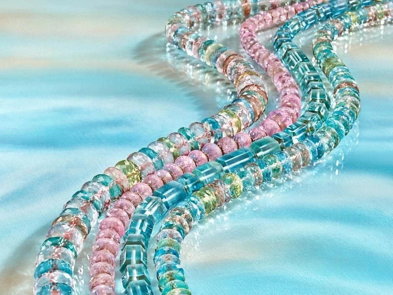 Beaded necklaces in shades of blue, pink, green, and clear on a shimmering blue surface, creating a vibrant jewelry display.