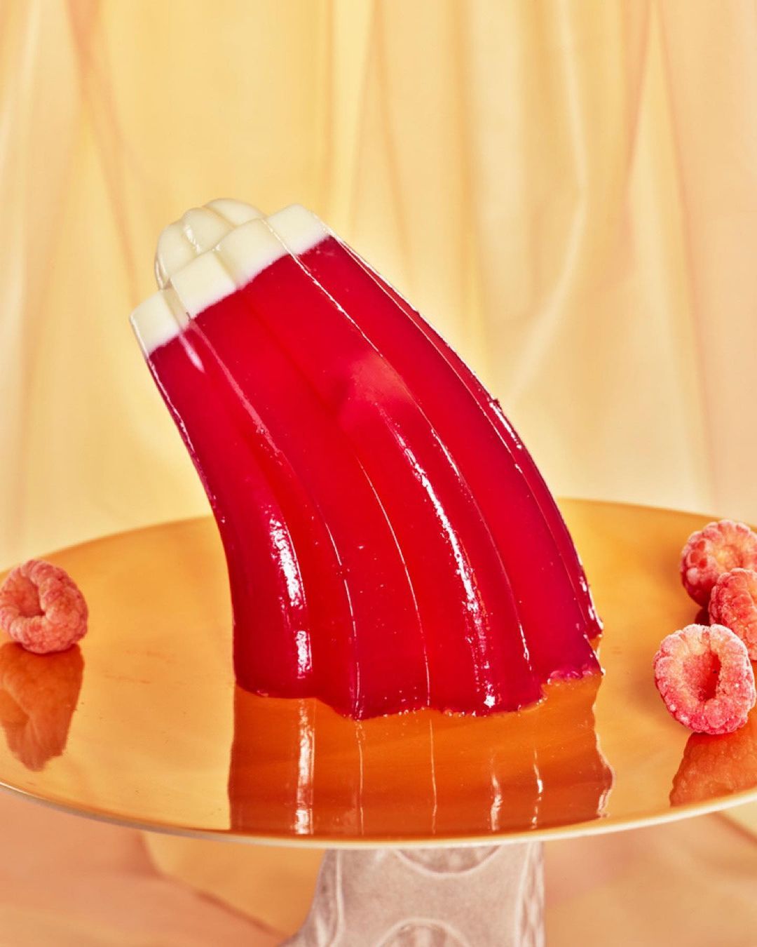 Red and orange jello dessert mold on a gold cake stand, with raspberries, against a cream and peach background.