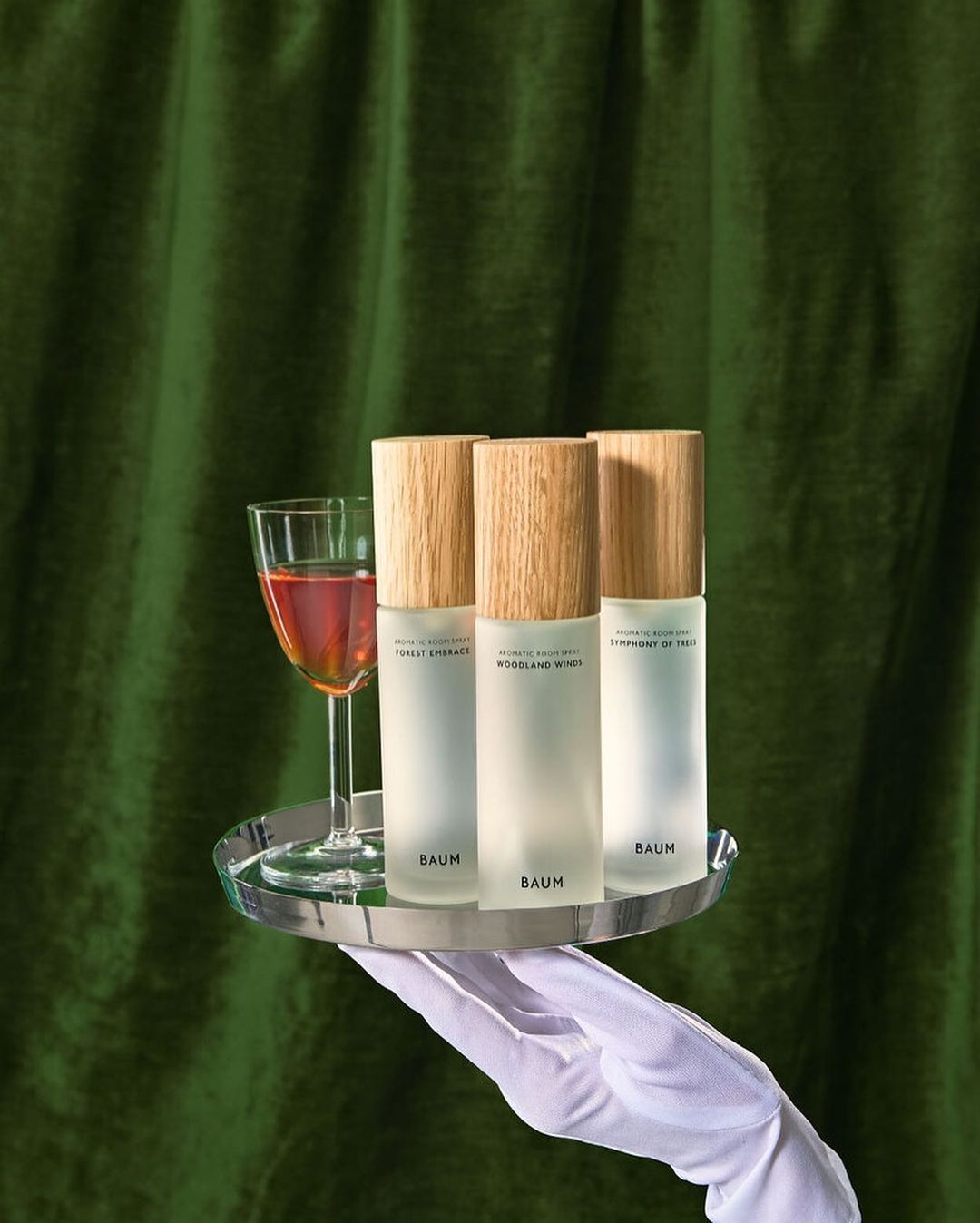 Still life product shot featuring three Baum skincare bottles with wooden caps on a silver tray with a drink, against a green velvet curtain.