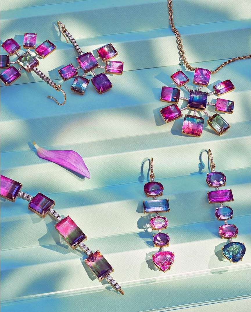 High-angle shot of jewelry including a necklace, bracelet, and earrings with pink, purple, blue, and clear gemstones displayed on light blue surface.