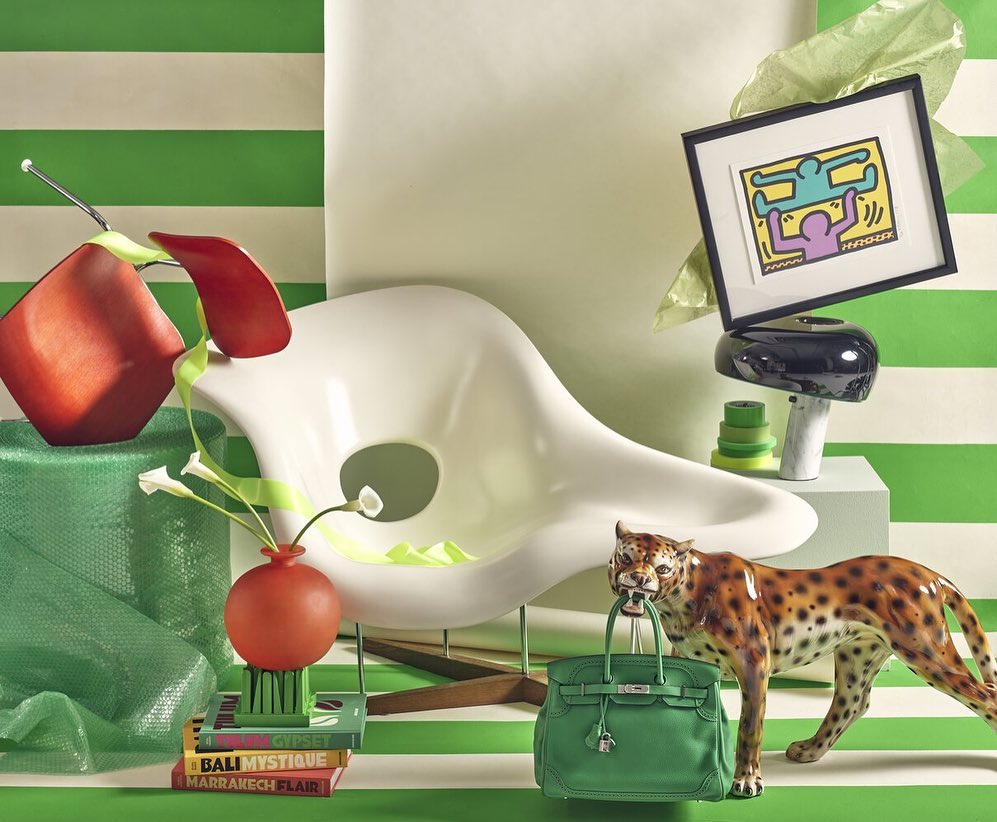 Still life of a white modern chair, framed art, ceramic leopard with green handbag, books, a lamp, and decorative objects, on a striped surface.