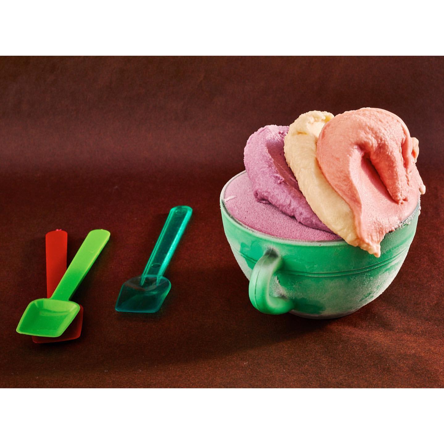 Close-up studio shot of a green cup filled with lavender, cream, and pink ice cream, with three colorful plastic shovels.