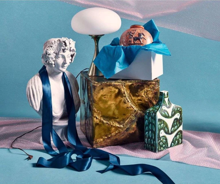 Still life arrangement featuring a bust draped with a blue ribbon, a lamp, two vases, and a metallic cube on a light blue backdrop.
