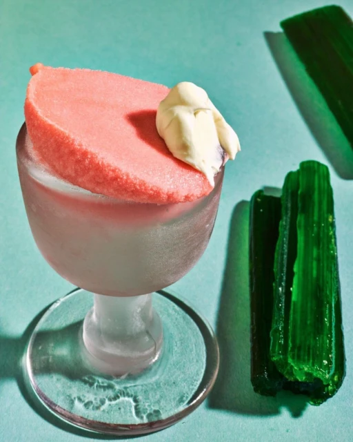 Still life of a stem glass topped with pink sorbet and cream, alongside green crystals on a light blue background. Food photography.