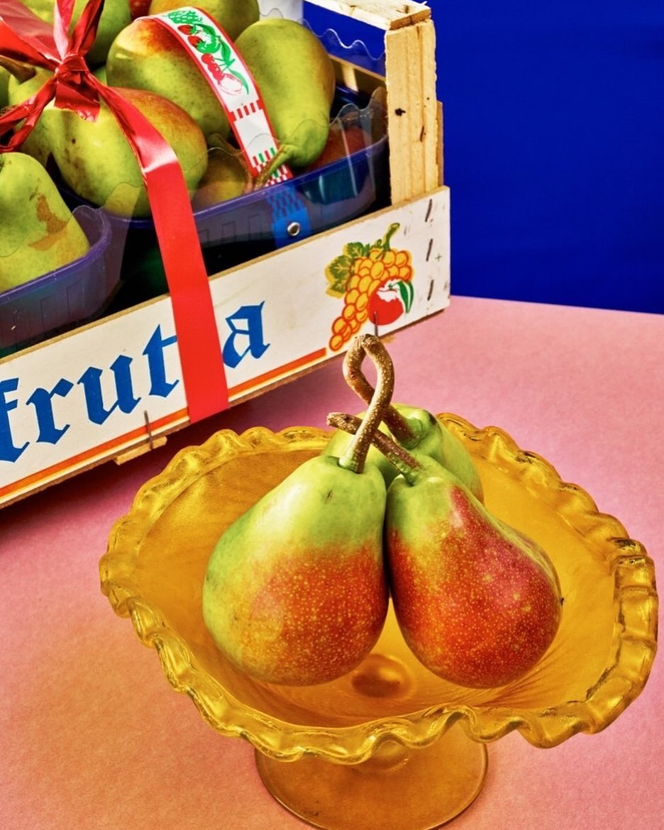 Close-up of a yellow pedestal bowl holding three pears, a crate filled with pears is behind it, against a pink and blue backdrop.