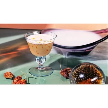 A coupe glass filled with a creamy tan drink sits on a reflective surface with dried peppers and a glass orb, with a bowl in the background.