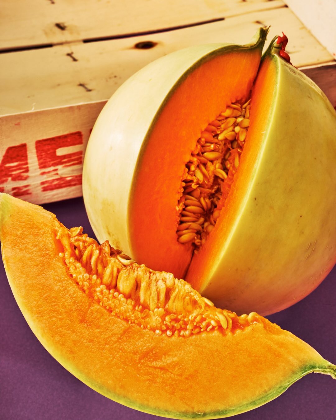 Close-up of a halved cantaloupe melon with seeds alongside a melon wedge, set against a wooden crate and a solid purple background.