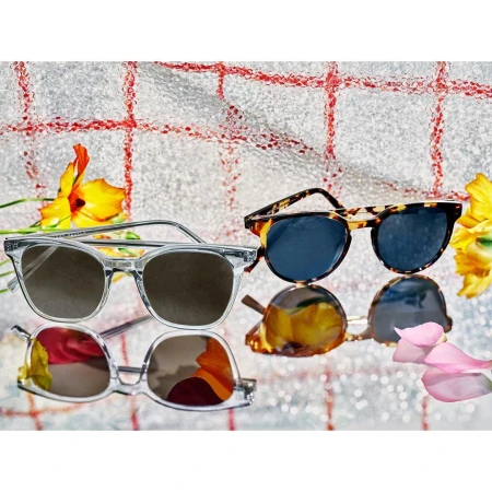 Two pairs of sunglasses, one clear-framed and one tortoise-shell, lay on a reflective surface with flowers and a checkered background.
