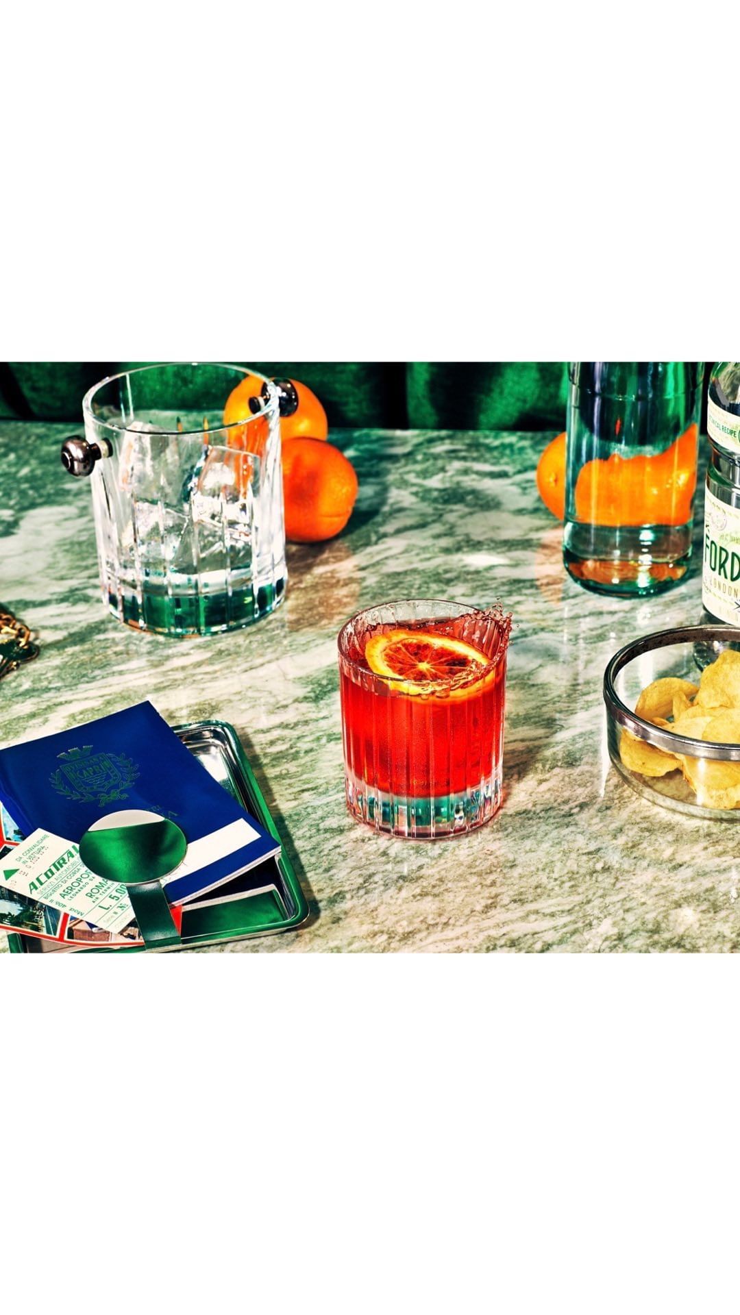 Cocktail with orange slice, spirits bottles, and assorted ingredients on a marble surface with ice bucket and passport.