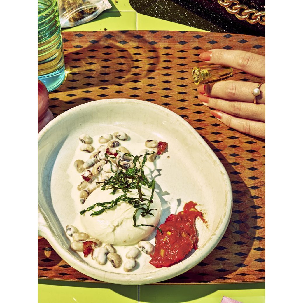 Burrata cheese sits on a plate with beans and tomato paste on a wooden surface next to a glass and hand wearing rings.