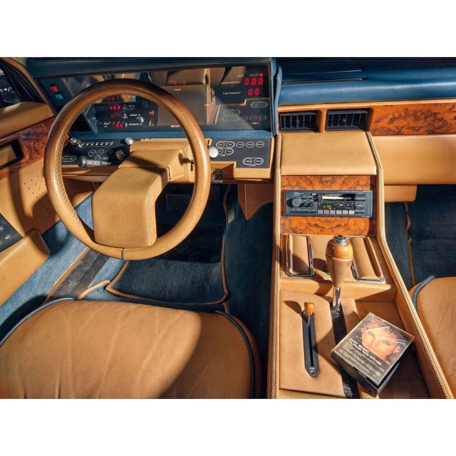 Retro car interior featuring a tan leather dashboard, blue carpeting, a wood steering wheel, and a vintage cassette tape.