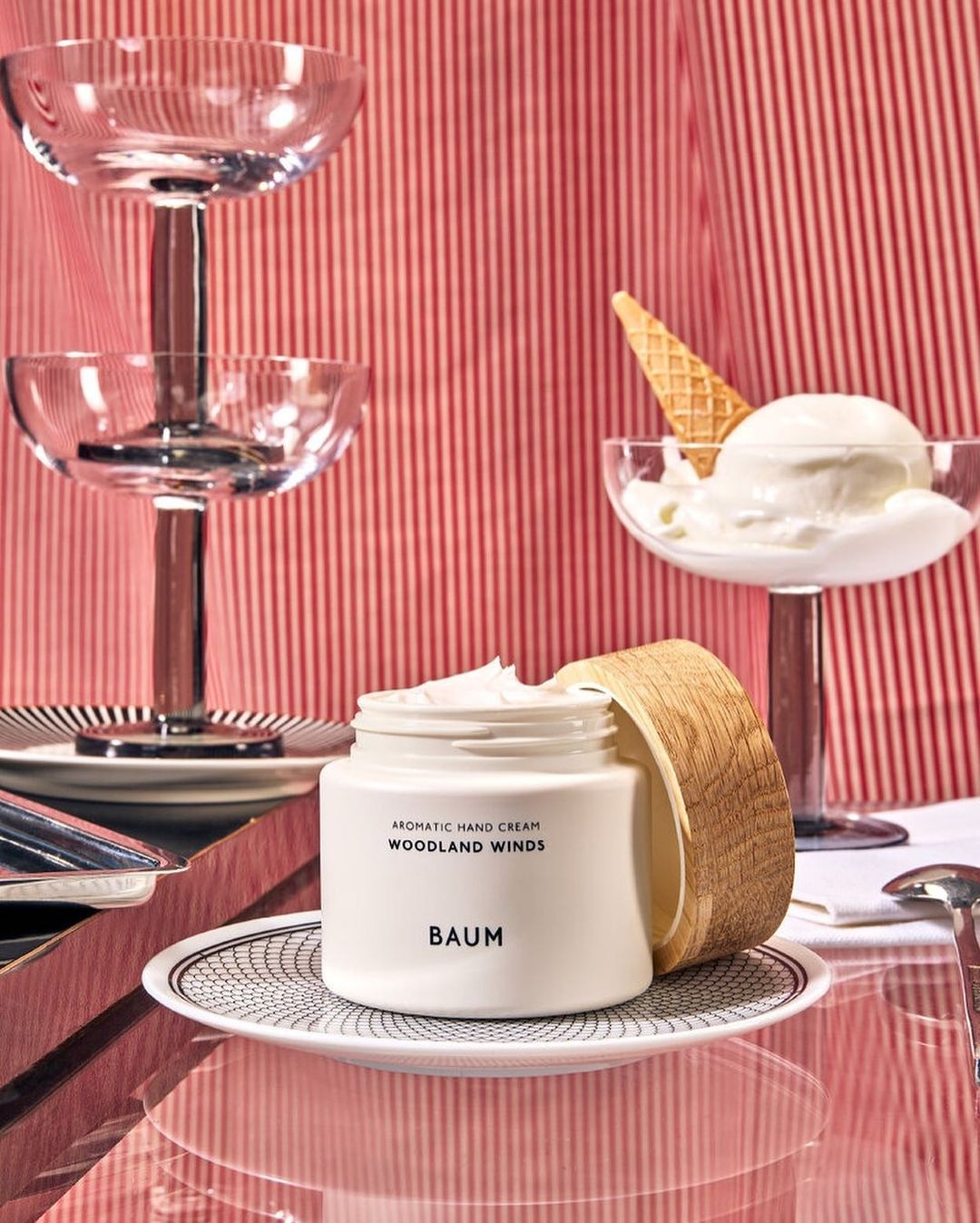 Still life of Baum hand cream jar on patterned plate with dessert glasses, ice cream cone, against red striped background.