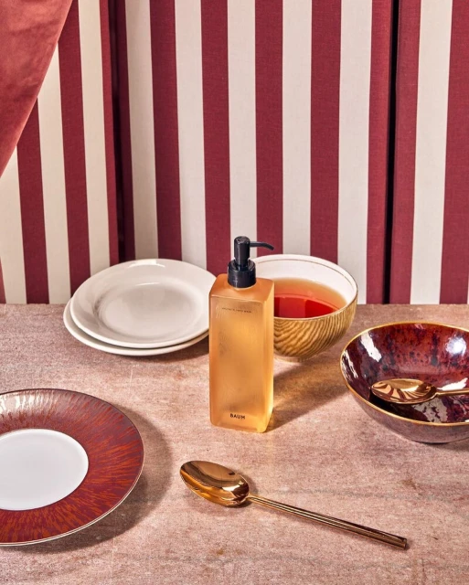 Still life shot in studio with gold and white dishes, spoons, and a pump bottle against a burgundy and white striped backdrop.