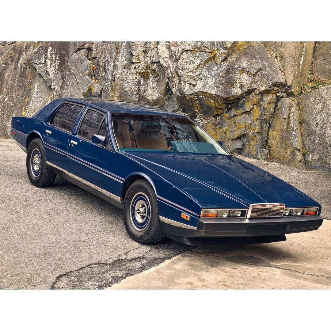 A dark blue vintage Aston Martin Lagonda sedan, parked on a concrete surface, against a rocky hillside backdrop, is captured in a medium shot.