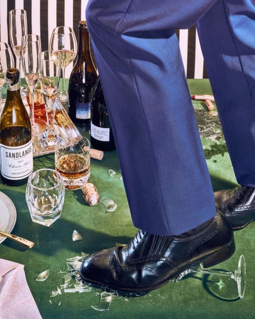 A man in a suit steps on a broken champagne glass on a table with bottles and glasses, creating a scene of luxury gone wrong.