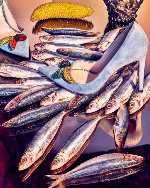 A pair of light blue stiletto heels artfully arranged with glistening fish and a shiny gold object on a reflective surface.