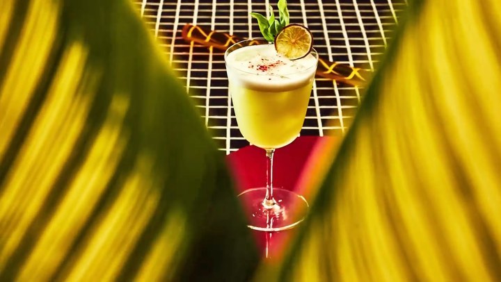 Cocktail in a stemmed glass with citrus garnish, set on a black and white checkered surface, accented by bold yellow foliage.