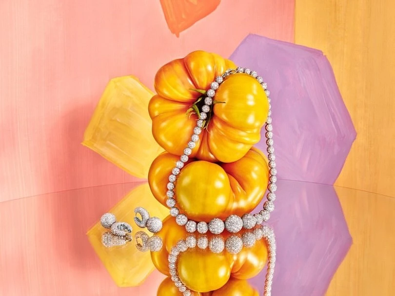 Yellow heirloom tomatoes stacked and adorned with a diamond necklace and earrings, reflected on a shiny surface, against a colourful backdrop.
