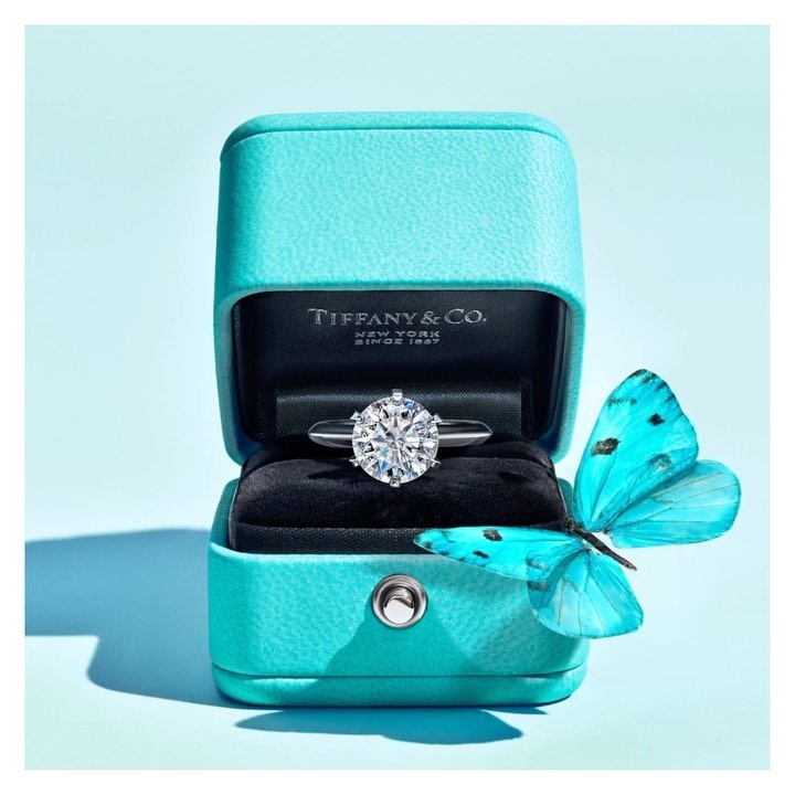 Close-up of a diamond solitaire ring inside a turquoise Tiffany & Co. box, with a turquoise butterfly resting against the box.