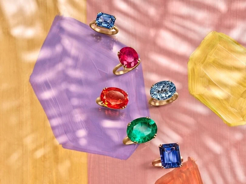 Overhead shot of gold rings with various coloured gemstones on a pastel painted surface, showcasing jewellery for fashion and beauty.