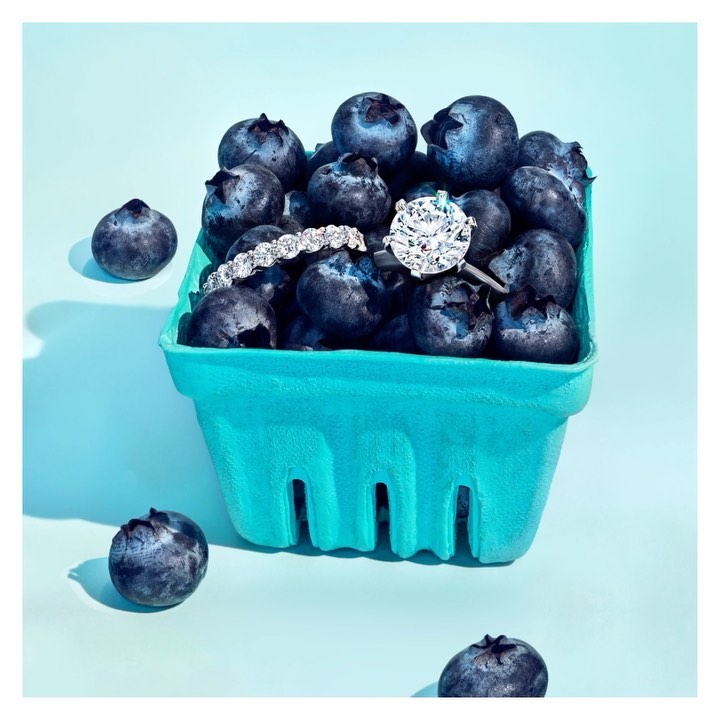 Close-up of a diamond ring and jeweled band nestled among fresh blueberries in a light-blue berry basket on a matching backdrop.