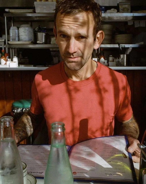Man in a red shirt sits at a diner table with water bottles and a menu, bathed in dappled sunlight from a window, indoor setting.