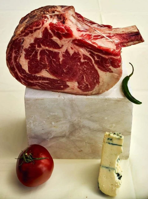 A raw tomahawk ribeye steak sits atop a marble block, accompanied by a tomato, blue cheese, and a green chili pepper in a food still life.
