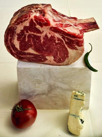 A raw tomahawk ribeye steak sits atop a marble block, accompanied by a tomato, blue cheese, and a green chili pepper in a food still life.