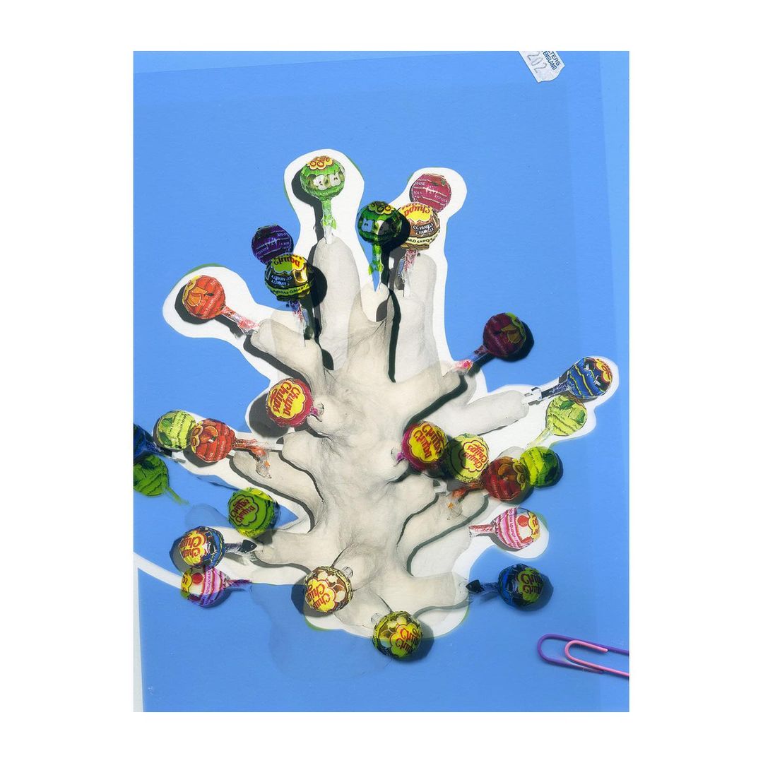 Surreal, overhead collage of Chupa Chups lollipops with wrappers arranged around a central paper shape on a light blue background.