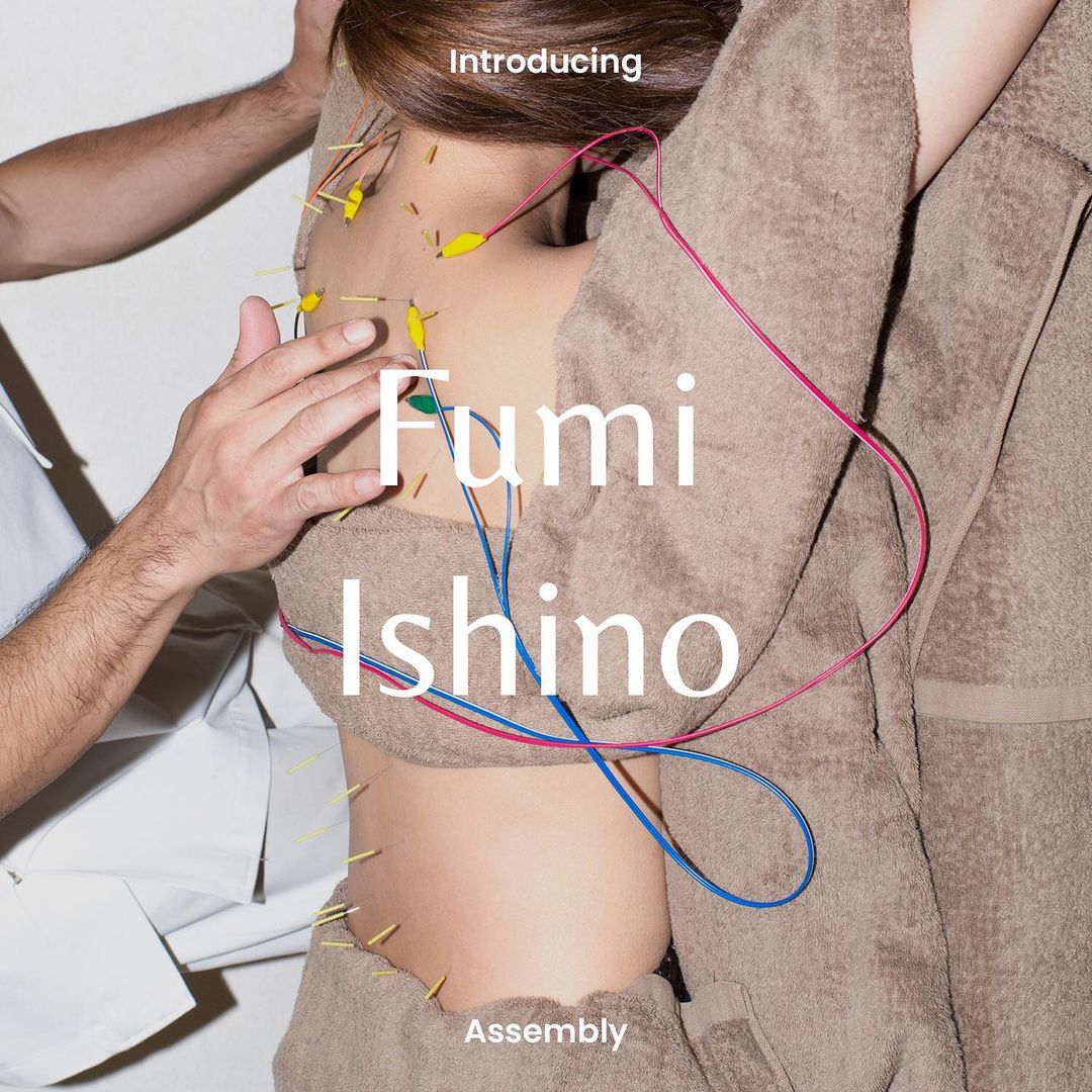 Close-up of a person's back and torso with multiple acupuncture needles, pink and blue cords, and another person's hands.