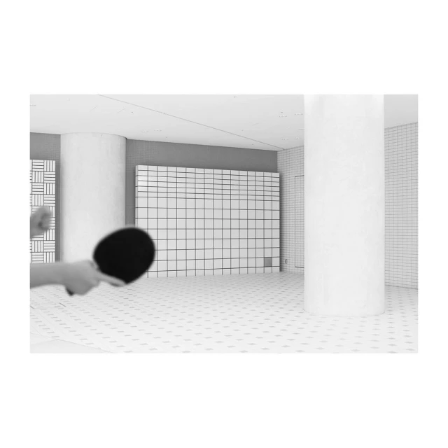 Monochrome shot of a person holding a ping pong paddle in an indoor tiled room with columns and a grid-like wall design.
