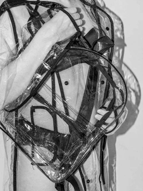 Person wearing a transparent raincoat and backpack, captured in black and white with studio lighting against a plain backdrop.