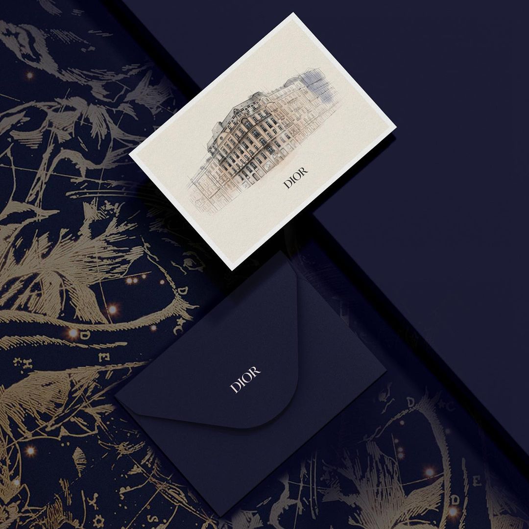 Stationery flat lay featuring a Dior building sketch card and a Dior envelope on a dark blue patterned surface, showcasing a luxury brand.