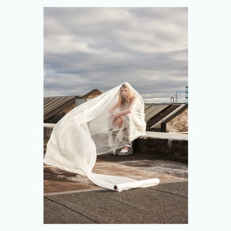 Blond woman in an embellished dress poses on a rooftop, draped in translucent plastic, with a cloudy sky backdrop and urban views.