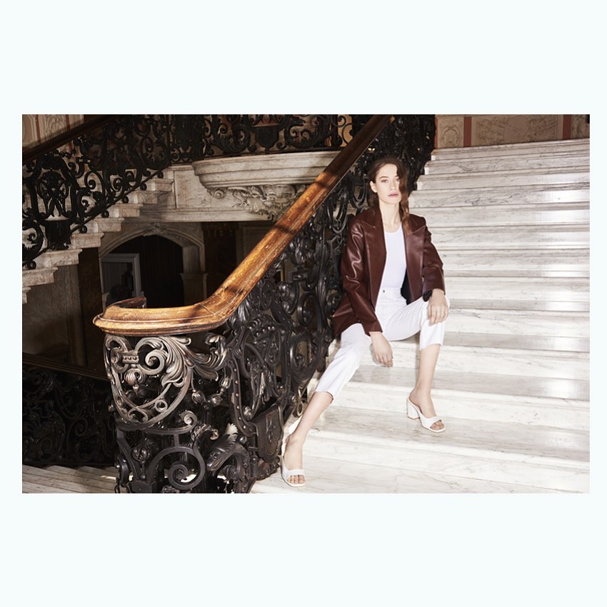 Adult female poses on staircase with ornamental iron railing wearing a dark red blazer, white top and capri pants, and light colored heels.