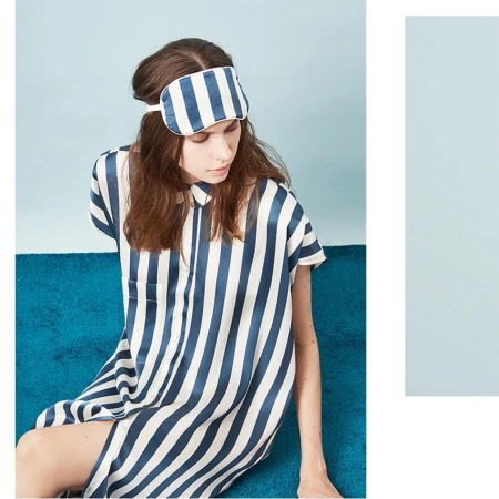 A woman in a striped blue and white dress and a matching sleep mask sits on a teal couch in front of a light blue wall.
