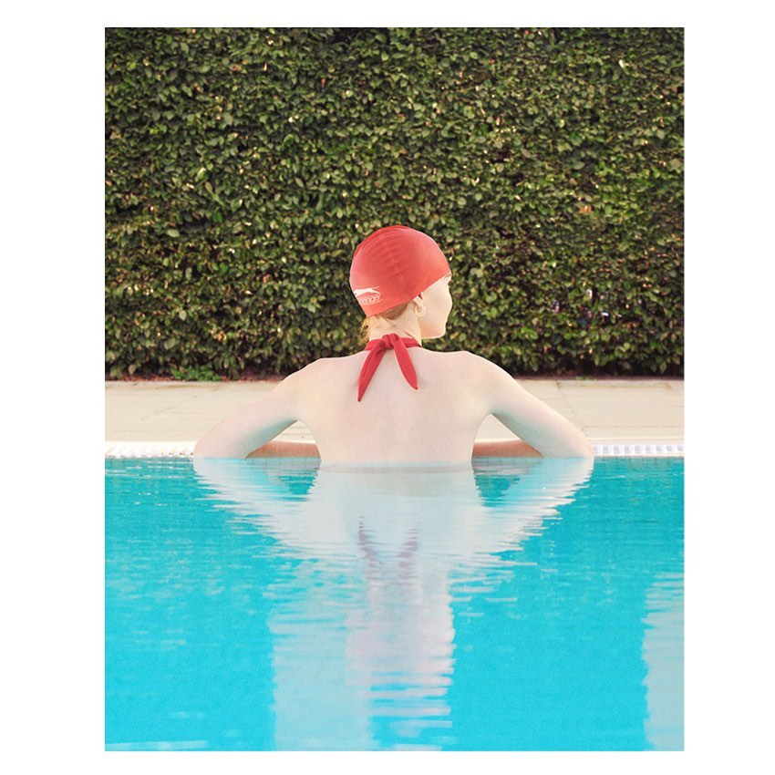 Woman in red swim cap at pool's edge, back to camera, looks at a wall of green foliage in bright, soft light with reflections.