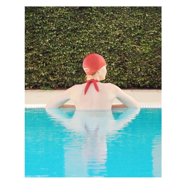 Woman in red swim cap at pool's edge, back to camera, looks at a wall of green foliage in bright, soft light with reflections.