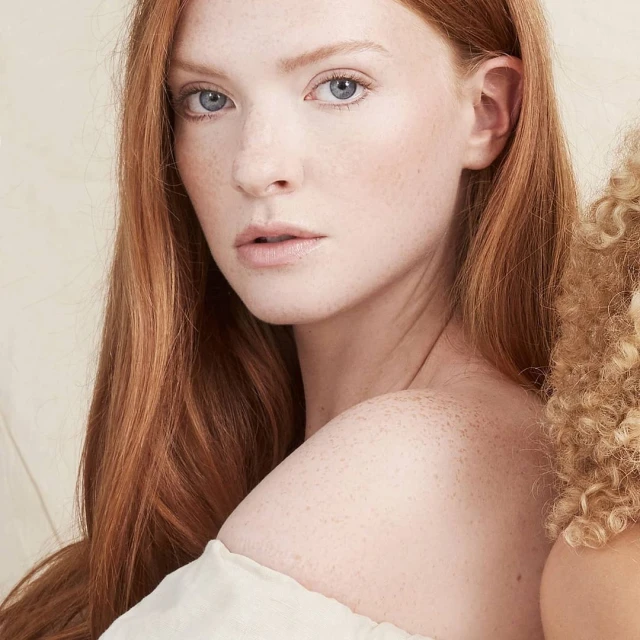 Close-up portrait of a fair-skinned woman with red hair and freckles, looking at the camera with a serious expression in a studio setting.