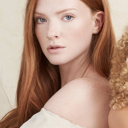 Close-up portrait of a fair-skinned woman with red hair and freckles, looking at the camera with a serious expression in a studio setting.