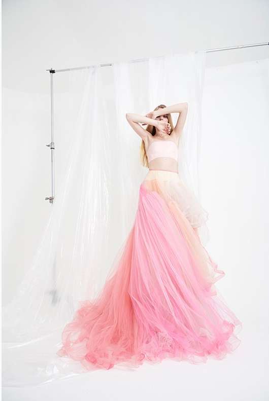 A woman in a flowing pink and peach dress stands in a photo studio, with her face obscured by her hands, creating an ethereal mood.