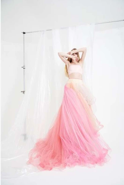 A woman in a flowing pink and peach dress stands in a photo studio, with her face obscured by her hands, creating an ethereal mood.