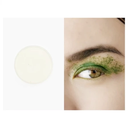 Close-up of a woman's eye with green glitter makeup alongside a container of white cream makeup on a white background.