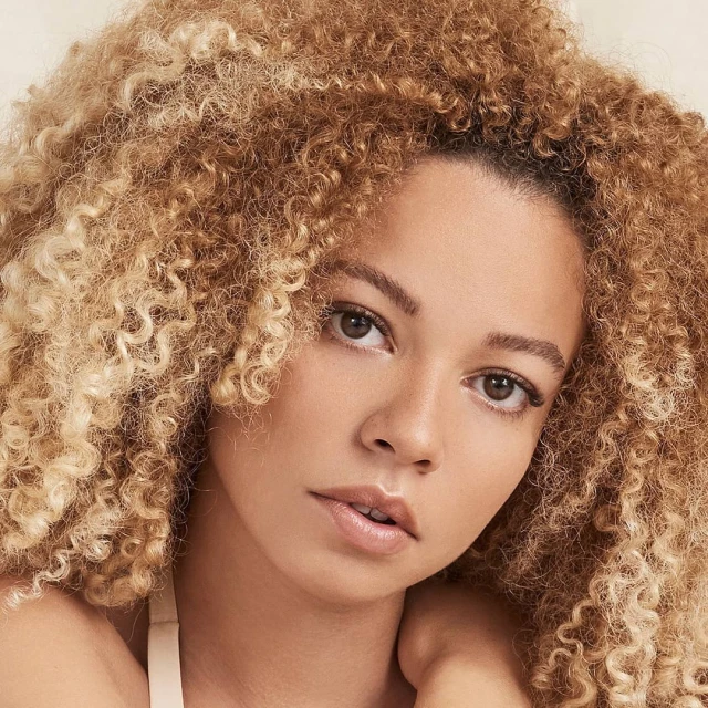 Close-up portrait of a woman with curly blonde hair looking at the camera with a neutral expression in a brightly lit studio.